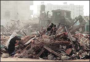 A woman retrieves what she can from the rubble of her home, while an armoured bulldozer follows its destructive path.